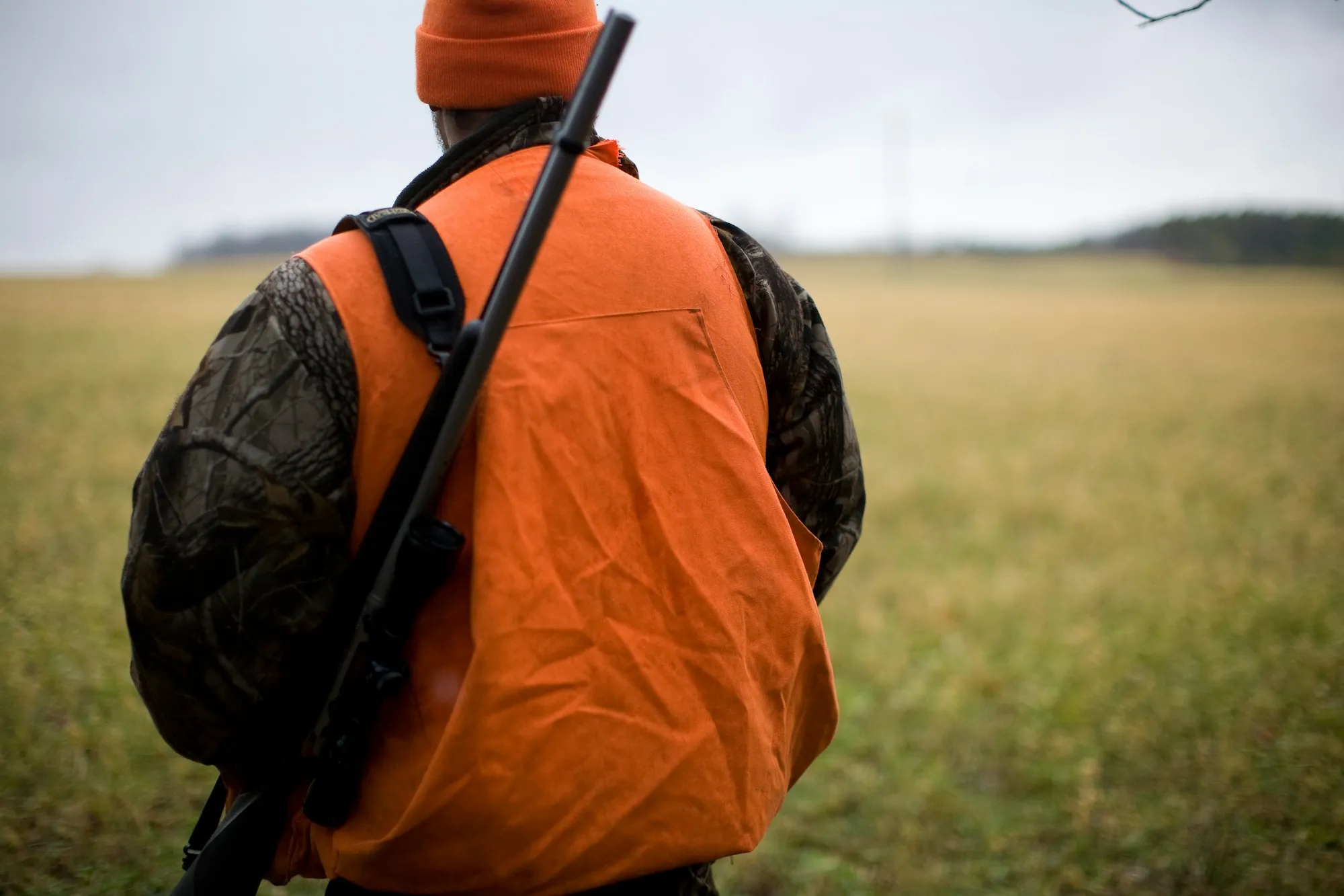 Jæger i orange vest set bagfra med våbnet i rem over skulderen ved en mark
